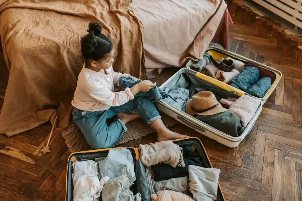 Child packing suitcase with clothes while sitting on floor near bed, preparing for travel adventure.
