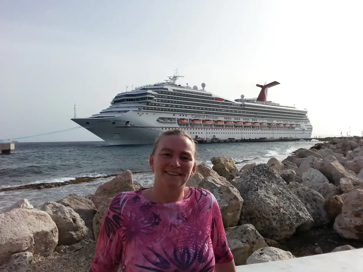 Smiling person in front of a large cruise ship by the rocky seaside, under a clear sky.