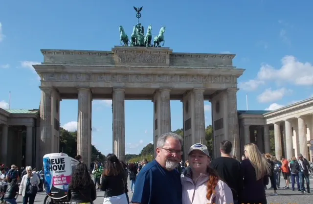 Couple posing at Brandenburg Gate, Berlin, with blue sky background and lively crowd.