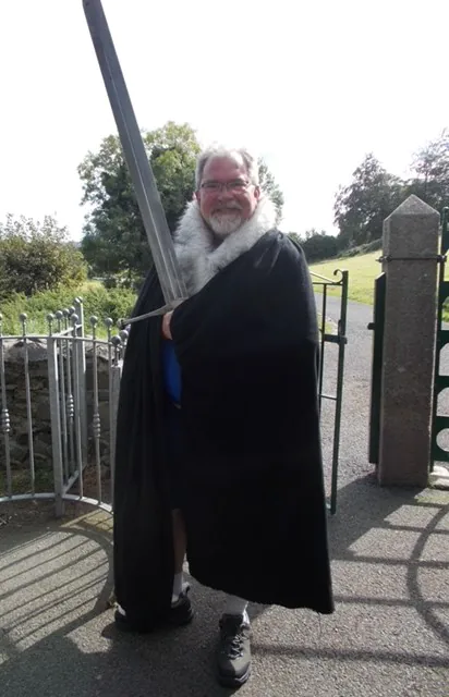 Man in dark cape with sword posing outdoors near gate, surrounded by greenery.