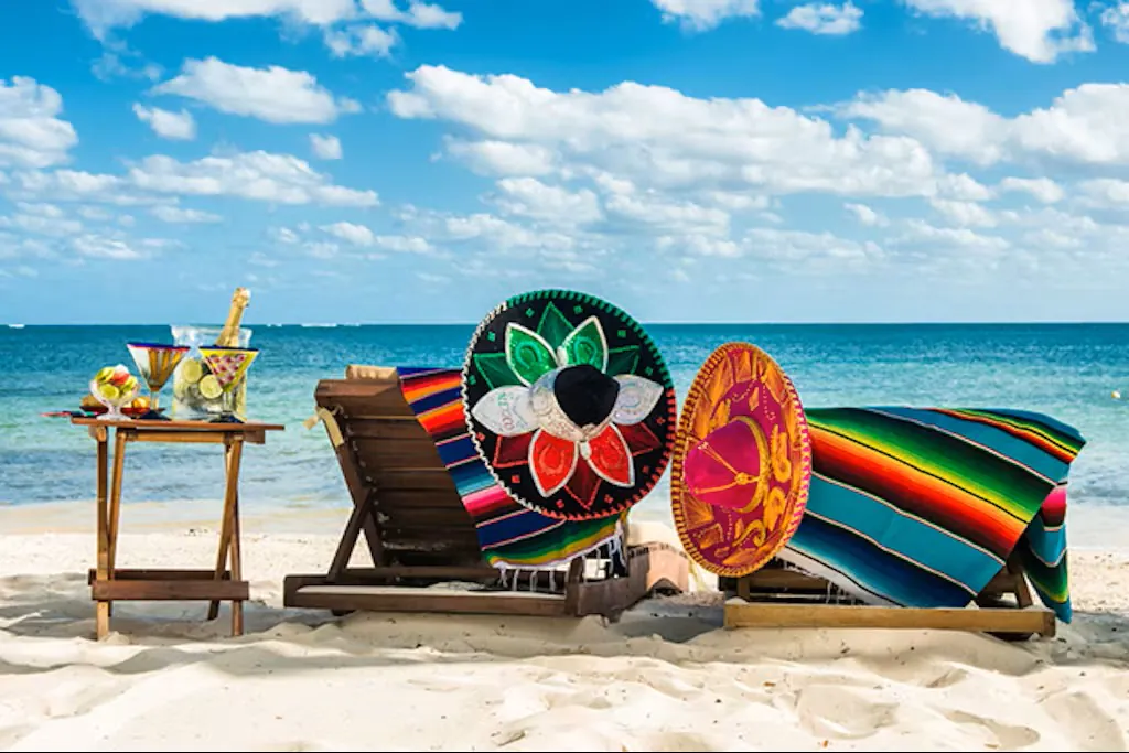 Colorful sombreros and serapes on beach chairs by the ocean, with cocktails on a side table under a vibrant blue sky.