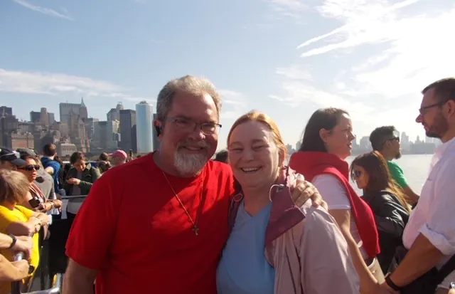 A smiling couple poses in front of a city skyline on a sunny day, surrounded by a crowd of people.