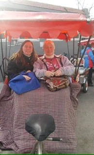 Couple enjoys a ride in a red canopy pedicab, bundled in blankets for warmth, with bags beside them.