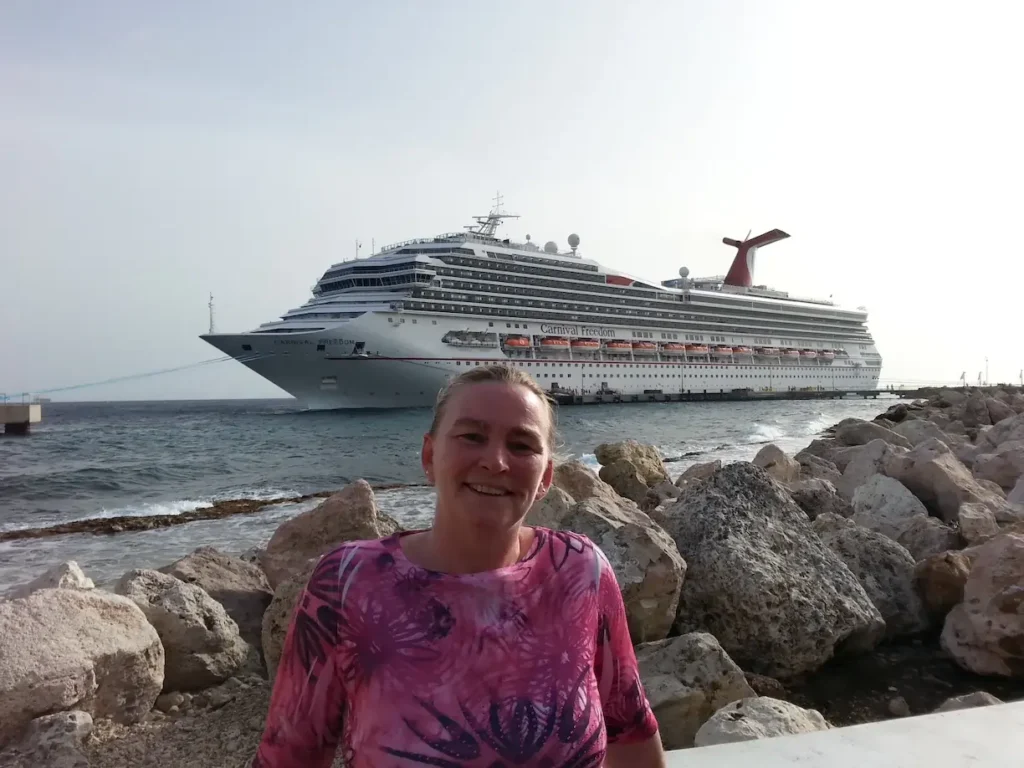 Smiling person in front of a large cruise ship docked at a rocky shore.