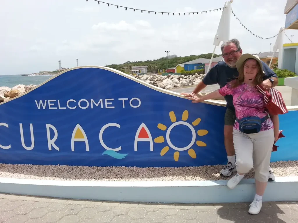 Couple smiling near Welcome to Curacao sign by the ocean, showcasing travel enthusiasm and tropical vibes.