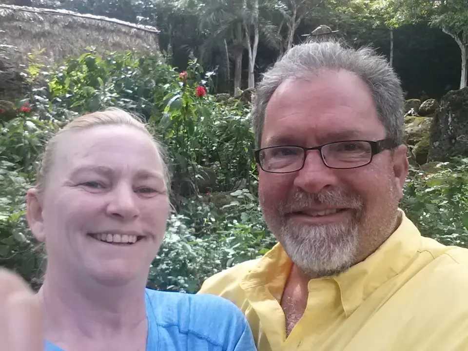Smiling couple taking a selfie in a lush garden with tropical greenery and rustic structures in the background.