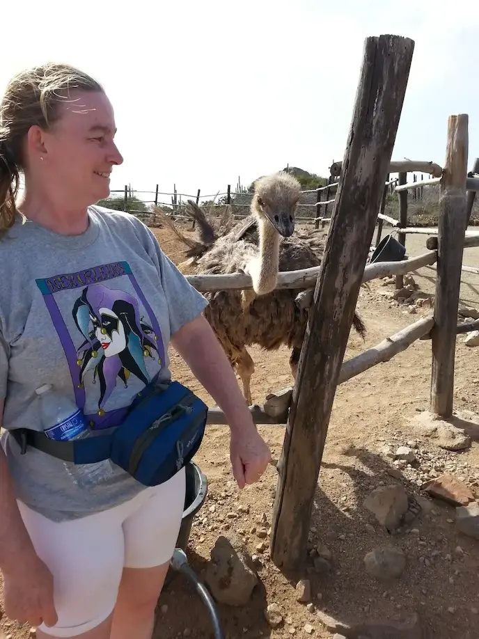 Woman smiling near an ostrich at a wooden fence in a sunny outdoor setting.