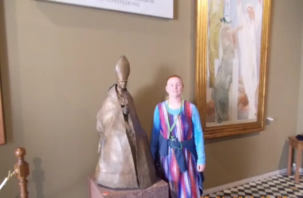 Woman standing next to a statue of a Pope in a gallery, colorful painting in the background.