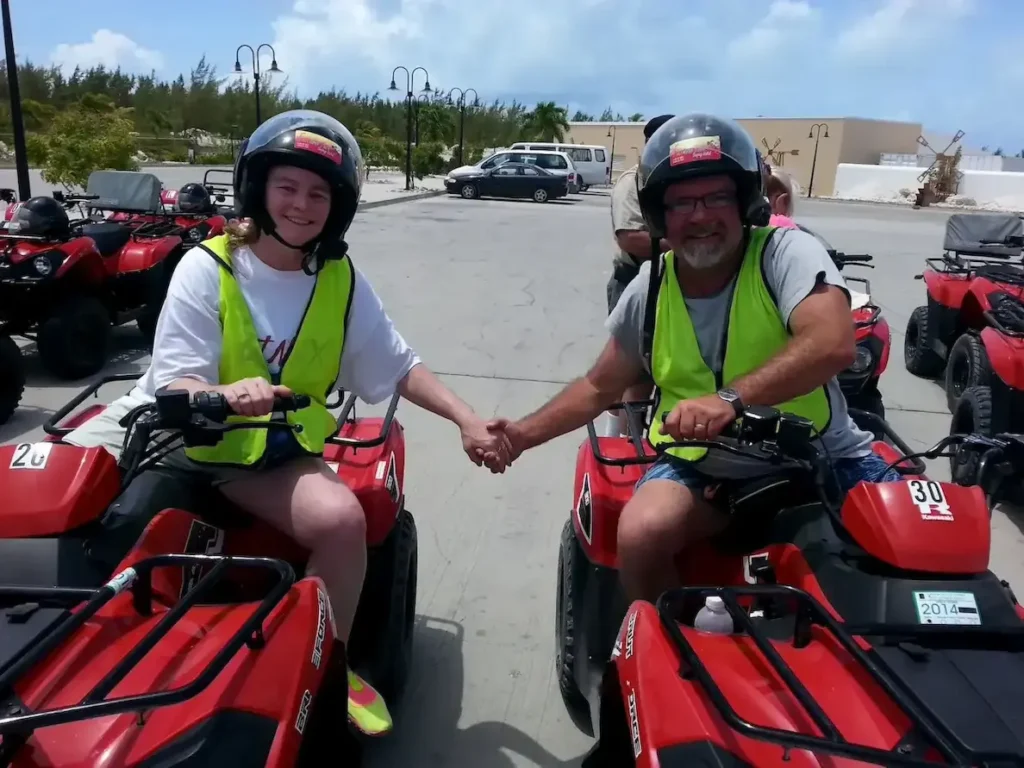 Two people in helmets holding hands on red ATVs, wearing neon vests, enjoy an outdoor adventure under a clear sky.