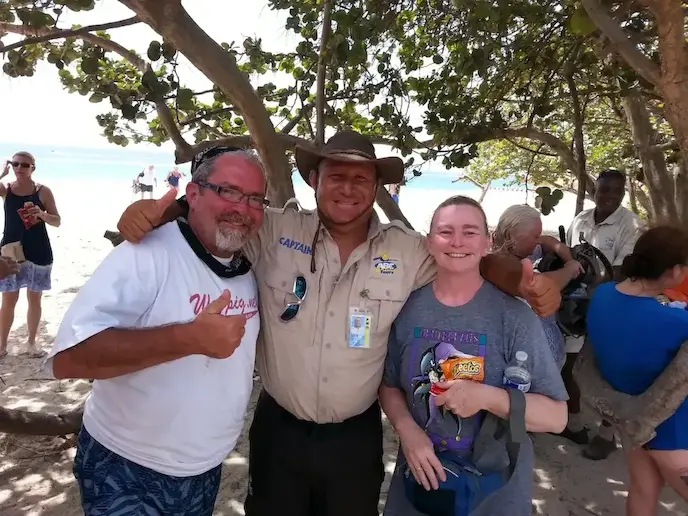 Group smiling on a beach with the sea in the background, trees providing shade on a sunny day.