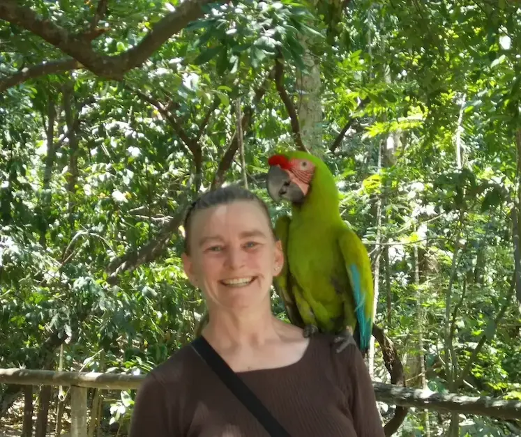 Smiling woman with a green parrot on her shoulder in a lush forest setting.
