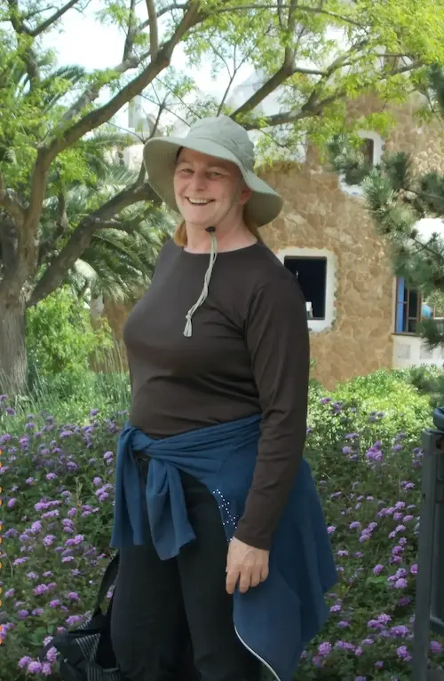 Woman in a hat smiling amidst flowers and trees in a sunny garden.