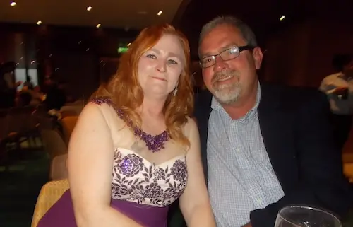 Couple smiling at an event, woman in purple dress, indoors with chairs in the background.