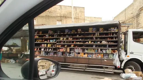 Mobile grocery store truck with stocked shelves in an urban area, viewed from a car window.