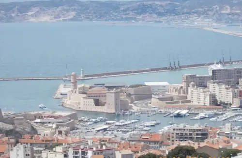 Aerial view of the Old Port of Marseille with boats, historic buildings, and a distant coastline on a clear day.