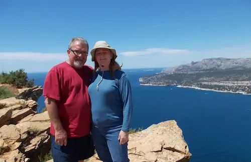 Man and woman standing on a cliff overlooking a blue ocean and distant hills under a clear sky.