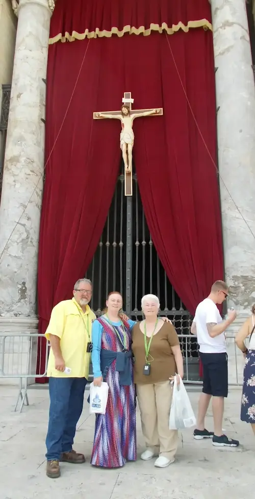 Three people stand in front of a church with a large crucifix and red curtain backdrop.